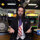 Billy Mitchell, wearing a black suit with an American flag tie, sites in front of two Donkey Kong barrels and a bunch of arcade cabinets.